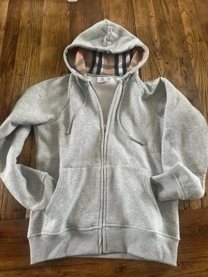 Grå zip hoodie  Burberry - Säljer en grå zip hoodie med klassiskt rutigt foder i huvan och dragsko. Hoodien har två fickor framtill och ribbade muddar vid ärmar och nederkant. Perfekt för en chill och stilren look. Stl M men passar S, Mått 44 bredd 66 lång, längde från armhålan 50. 