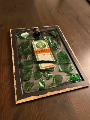 Krossad jäger tavla - Unikt konstverk med krossade gröna glasbitar och en Jägermeister-flaska inramad i svart ram. Flaskans etikett och en miniatyrflaska är synliga bland glasskärvorna, vilket ger en cool och kreativ look. Perfekt som dekoration för hemmabaren eller som samtalsämne.