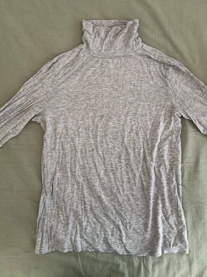 Ljusgrå polotopp i mjukt material - Säljer en ljusgrå polotopp med lång ärm och hög krage. Toppen är tillverkad i ett mjukt och stretchigt material som sitter skönt mot kroppen. Perfekt att styla lager på lager eller bära som den är för en clean look.