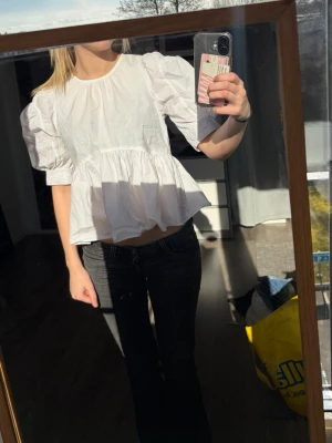 Vit puffärmad blus med volang - Säljer en vit blus med puffiga trekvartsärmar och volang nedtill. Blusen har rund halsringning och knäppning i ryggen. Tillverkad i ett lätt bomullsliknande material som ger en luftig känsla. Perfekt för en trendig och avslappnad look.