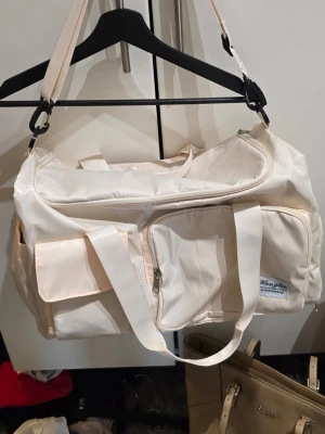 Beige weekendbag ny - Rymlig beige weekendbag med flera smarta fack och dragkedjor. Väskan har både axelrem och handtag, samt praktiska ytterfickor för enkel organisering. Tillverkad i slitstarkt syntetmaterial med stilren design.