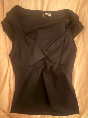 söt svart topp  - Snygg svart omlottopp från H&M med korta ärmar och v-ringad hals. Toppen har en figurnära passform och är tillverkad i mjukt stretchigt material, perfekt för att lyfta vilken outfit som helst.