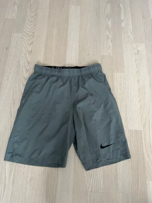 Nike Träningsshorts - Snygga grå träningsshorts från Nike med elastisk midja och diskret svart logga på vänster ben. Shortsen har en enkel och sportig design med två sidofickor och är tillverkade i ett lätt syntetmaterial som passar perfekt för aktiva dagar eller till sommaren. Även om storleken är S så kan den passa till M