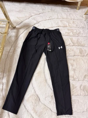 Svarta mjukisbyxor från Under Armour - Snygga svarta mjukisbyxor från Under Armour med resår i midjan och dragsko. Byxorna har raka ben, sidofickor med dragkedja och en vit logga på låret. Perfekta för chill eller träning, tillverkade i ett mjukt och lätt material. Passar som S
