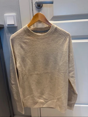 Beige stickad tröja i ullmix - En enkel och stilren beige stickad tröja med rund halsringning och ribbade muddar vid ärmslut och nederkant. Tröjan har en rak passform och är perfekt för lager på lager. Materialet känns mjukt och värmande, troligen en ullmix. Storlek L men passar M
