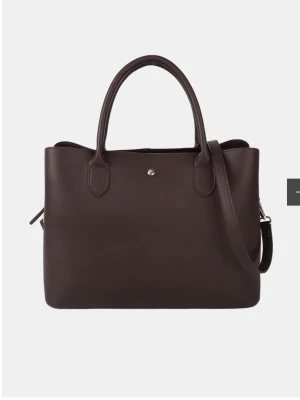 Mörkbrun handväska i konstläder - Stilren mörkbrun handväska i slätt konstläder med både handtag och justerbar axelrem. Väskan har en enkel, rektangulär form och en liten guldfärgad knappdetalj framtill. Perfekt för dig som gillar minimalistisk design och vill ha plats för allt du behöver. Den har även ett stort fack för dator.