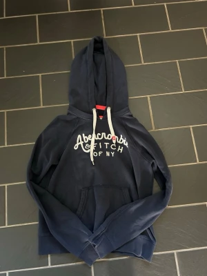 Mörkblå hoodie Abercrombie & Fitch - Mörkblå hoodie från Abercrombie & Fitch med vit broderad logga och text 'OF NY' på bröstet. Klassisk känguruficka framtill, vita dragsnören i huvan och ribbade muddar. Perfekt för en avslappnad och trendig stil. Sitter mer som xs eller s i storlek