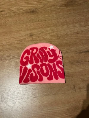 Rosa mössa Gravity  Visions spindelnät - Säljer en rosa mössa med coolt röd texttryck 'Gravity Visions' och vita stjärnor på framsidan samt ett spindelnätsmönster på baksidan. Mössan är stickad i mjukt material och har en avslappnad passform. Perfekt för dig som gillar unik streetwear och grafiska detaljer.