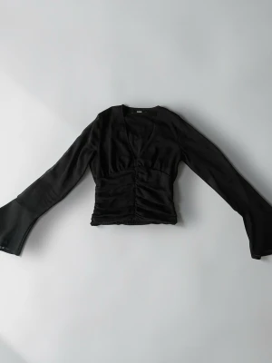 Svart omlottopp med v-ringning - Trendig svart omlottopp med v-ringning och långa ärmar. Toppen har snygg rynkning framtill och är croppad i modellen. Tillverkad i ett mjukt, lätt material som ger en elegant look.