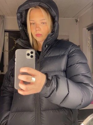 Svart pufferjacka från Peak Performance - Säljer en svart pufferjacka från Peak Performance med huva och dragkedja framtill. Jackan har ett stilrent och sportigt utseende med quiltade paneler och logotyp på bröstet. Perfekt för kalla vinterdagar och håller dig varm och bekväm. Köpt förra året så skicket är som nytt.