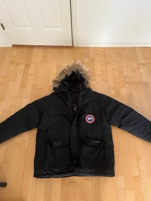 Svart jacka från Canada Goose - Säljer en svart jacka från Canada Goose med klassisk patch på bröstet och stor huva med fuskpäls. Jackan har flera stora fickor framtill och dragkedja med vindslå. Perfekt för kalla vinterdagar och riktigt varm. Storlek M herr! Skriv till mig vid intresse eller frågor! 🤗