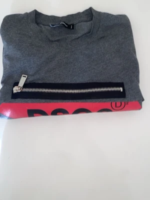 Grå DSQUARED2 topp med dragkedja - Cool grå topp från DSQUARED2 med stor röd logga och unik silverfärgad dragkedja framtill. Rund halsringning och rak passform. Perfekt för dig som gillar streetstyle och vill sticka ut med detaljer.