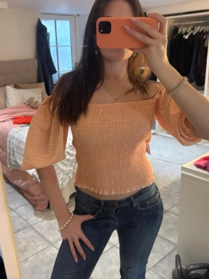 Corall offshoulder blus med puffärm - Superfin orange/corall offshoulder blus med rutigt mönster och smockad kropp. Blusen har vida puffärmar och croppad passform, perfekt för en trendig look. Lätt och luftig, passar perfekt till jeans eller kjol.