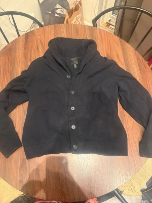 Svart kofta med sjalkrage och knappar - Stilren svart kofta med sjalkrage och knäppning framtill. Koftan har långa ärmar, ribbade muddar och en mjuk, stickad känsla. Perfekt för lager på lager och enkel att matcha med olika outfits.