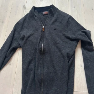 Mörkgrå ziptröja från morris - Mörkgrå ziptröja från Morris med hög krage och hel dragkedja framtill. Tröjan har en diskret logga på bröstet och ribbade muddar vid ärmslut och nederkant. Perfekt för lager på lager och en clean look.