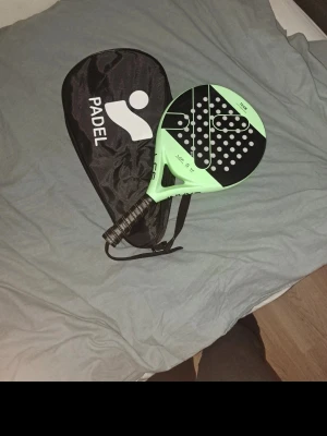 Grön och svart padelracket med fodral - Snygg padelracket i grön och svart design med matchande fodral. Racketen har perforerat blad och greppvänligt handtag, perfekt för dig som vill ta ditt spel till nästa nivå. Fodralet har bärrem och tydlig logga. Passar både träning och match.