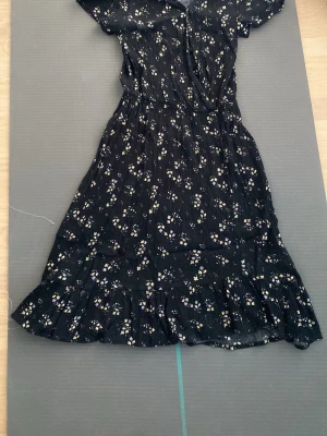 Svart blommig långklänning - Svart långklänning med små vita och gula blommor, volangkant nertill och korta ärmar. Klänningen har en feminin siluett med markerad midja och v-ringning. Perfekt för sommarens alla tillfällen.