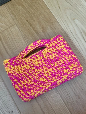 Handvirkad kuvertväska i rosa och gul - Unik handvirkad kuvertväska i starka färger, mixad i rosa och gul. Väskan har inbyggt handtag och är gjord av tjockt garn som ger en cool och trendig look. Perfekt accessoar för dig som vill sticka ut och addera färg till din outfit.