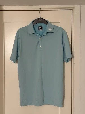 Helt ny golfpiké från FootJoy - Snygg ljusblå pikétröja från FootJoy med klassisk krage och två knappar framtill. Tillverkad i mjukt och lätt polyestermaterial som andas. Kortärmad modell med diskret logga på kragen och ryggen. Perfekt för en sportig och avslappnad stil. Köpt ny förra säsongen 