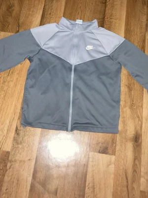Grå ziptröja från Nike Dri-Fit - Snygg grå ziptröja från Nike med Dri-Fit-material. Tröjan har två olika grå nyanser, dragkedja framtill och en liten vit Nike-logga på bröstet. Perfekt för träning eller chill. Tillverkad i ett mjukt och funktionellt material.
