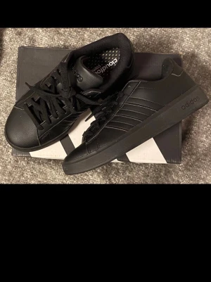 Svarta Adidas sneakers i läder - Oanvända Snygga helsvarta sneakers från Adidas med klassiska tre ränder på sidan. Skorna är låga och har ovandel i slätt läder, svart sula och svarta skosnören. Perfekta för dig som gillar en clean och stilren look. 