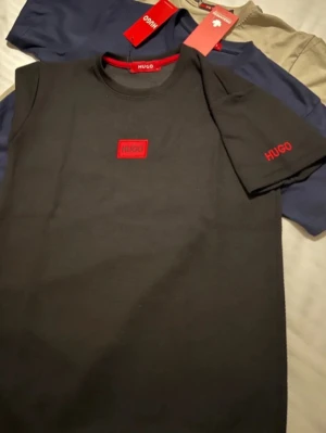 Svart t-shirt från HUGO med röd logga - Snygg svart t-shirt från HUGO med röd logga på bröstet och ärmen. Klassisk rund halsringning och normal passform. Perfekt basplagg med stilren design och diskreta detaljer. Tillverkad i mjuk bomull för skön känsla.