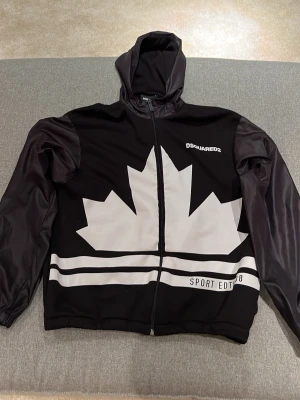Svart hoodie från Dsquared2 - Säljer en svart jacka/hoodie från Dsquared2 med stor vit lönnlövsgrafik på framsidan. Jackan har dragkedja, huva och vita ränder nedtill. Perfekt för dig som gillar streetwere. Skick 10/10 som ny. Storlek 14 år men som xs/s