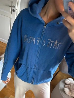 Blå hoodie med strasstext och figur - Blå zip-hoodie med huva och fickor framtill. På bröstet står det 'STATE OF MIND' i strass och på ryggen finns en stickman-figur i strass. Hoodien har avslappnad passform och är gjord i mjukt bomullsmaterial. Perfekt för en chill och trendig look. (UNIK UPPLAGA FINNS EJ ATT KÖPA LÄNGRE)