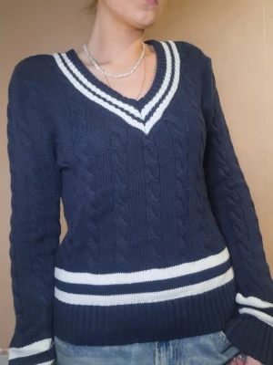 Marinblå kabelstickad tröja - Mörkblå kabelstickad tröja med vita ränder vid ärmslut, midja och krage. Tröjan har en klassisk, sportig look med v-ringad krage och ribbstickade muddar. Perfekt för dig som gillar preppy stil och vill ha något snyggt och bekvämt.