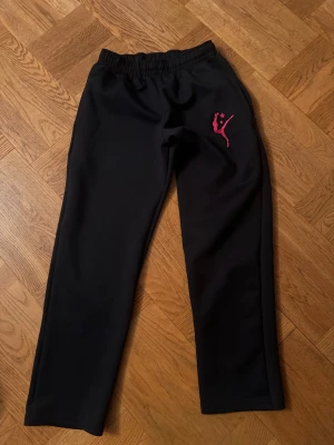 Svarta mjukisbyxor med dansmotiv - Säljer ett par svarta mjukisbyxor med resår i midjan och ett rosa broderat dansmotiv på ena benet. Byxorna har raka ben och är gjorda i ett mjukt och bekvämt material, perfekta för chill eller träning. Passar dig som gillar enkel och sportig stil.