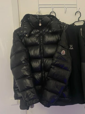 Moncler maya  - Snygg svart dunjacka från Moncler med glansig finish och klassisk puffer-design. Jackan har hög krage, dragkedja framtill och Moncler-logga på ärmen. Perfekt för kalla vinterdagar och riktigt trendig look.