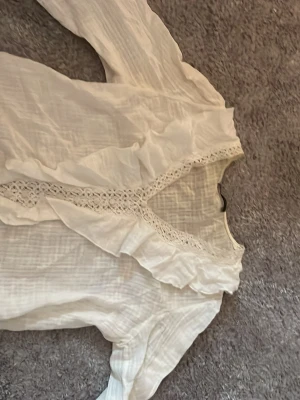 Vit blus med spets och volanger - Säljer en romantisk vit blus med långa ärmar, fina volangdetaljer och spets längs ringningen. Blusen har en luftig och lätt struktur, perfekt för en trendig och feminin look. Materialet känns mjukt och svalt mot huden.