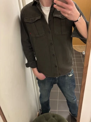 Mörkgrön overshirt med fickor - Stilren mörkgrön overshirt med två bröstfickor med lock och knapp, klassisk krage och knäppning framtill. Skön och avslappnad passform, perfekt att bära över en t-shirt. Materialet ser ut att vara kraftig bomull. Snygg detalj med liten röd prick på ena fickan.