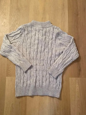 Ljusgrå kabelstickad tröja Polo Ralph Lauren - Ljusgrå kabelstickad tröja från Polo Ralph Lauren med halv dragkedja och ribbade muddar. Tröjan har en klassisk polokrage och den ikoniska broderade loggan på bröstet. Perfekt för lager på lager och riktigt snygg till jeans.