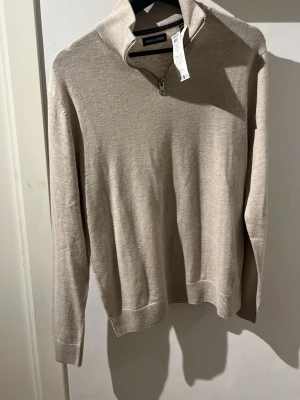 Beige half zip tröja Jack & Jones - Snygg beige half zip från Jack & Jones med halv dragkedja vid halsen. Tröjan har ribbade muddar vid ärmslut och nederkant, och är perfekt för lager på lager. Enkel och stilren design som passar till det mesta. Helt ny.