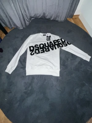 Vit sweatshirt från Dsquared2 - Säljer en vit sweatshirt från Dsquared2 med svart, stor logga tryckt diagonalt över bröstet. Tröjan har rund halsringning, långa ärmar och ribbade muddar vid ärmslut och nederkant. Tillverkad i mjukt bomullsmaterial för en skön känsla.