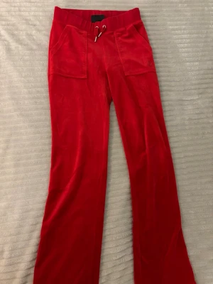 Röda velourbyxor Juicy Couture - Snygga röda mjukisbyxor från Juicy Couture i mjuk velour. Byxorna har bred resår i midjan med dragsko och stora fickor fram. Klassisk logga broderad på ena fickan. Perfekta för en chill och trendig look.