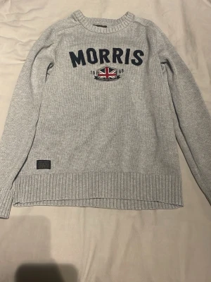 Ljusgrå stickad tröja Morris - Ljusgrå stickad tröja från Morris med rund halsringning och ribbade muddar. Framtill står det 'MORRIS' i mörkblått med ett brittiskt flaggmärke och årtalet 1968. Tröjan har en klassisk passform.