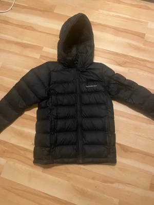 Svart dunjacka Peak Performance - Säljer en svart dunjacka från Peak Performance med huva och dragkedja framtill. Jackan har en puffig design och är perfekt för kalla vinterdagar. Märkeslogga broderad på bröstet. Klassisk och stilren modell som håller dig varm.