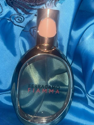 Ferragamo Fiamma parfym - Ferragamo Fiamma parfym i en elegant oval glasflaska med silverfärgade detaljer och en modern, stilren design. Flaskan har en transparent kropp med rosa text och ett unikt lock med beige detalj. Perfekt för dig som gillar exklusiva dofter. 100ml har ej kvar kartongen men ny köpt!! Använd 1gång inköps pris 1500kr