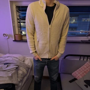 Beige ziptröja- indicode - Säljer en beige ziptröja med hög krage och ribbade muddar. Tröjan har ett diskret rutmönster i materialet och dragkedja framtill. Perfekt att slänga på sig över en t-shirt för en avslappnad look. Tröjan är i storlek M och är i bra skick!