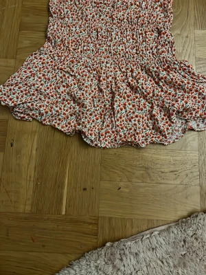 Blommig volangkjol från Pacific Republic - Supersöt kort kjol från Pacific Republic med blommigt mönster i rött och vitt. Kjolen har smock i midjan och volangkant nedtill.