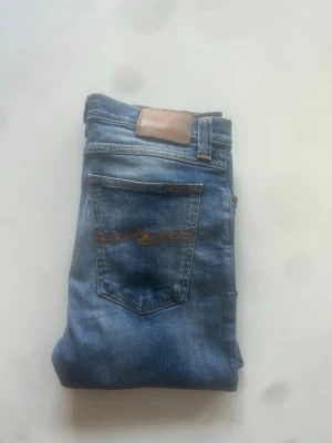 Blå jeans från Nudie Jeans - Säljer ett par klassiska blå jeans från Nudie Jeans med snygg slitning och kontrastsömmar. Modellen har fem fickor, normal midja och smal passform. Tillverkade i mjukt denimtyg med Nudie-logga på bakfickan.
