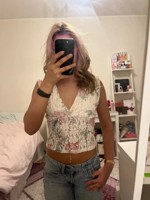 Blommig croppad spetsblus - Säljer en vit croppad blus med blommigt mönster och spetsdetaljer. Toppen har breda axelband, v-ringning och är ärmlös. Perfekt för dig som gillar romantiska och trendiga plagg med en feminin touch.