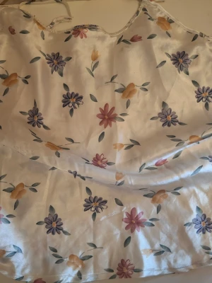 Blommigt nattlinne med tunna axelband - Sidenliknande nattlinne i ljus cremefärg med blommigt mönster i lila, gult och rosa. Tunna axelband och lös passform ger en avslappnad känsla. Perfekt för mysiga kvällar hemma.