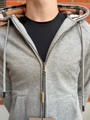 Grå zip hoodie från Burberry - Snygg grå hoodie med dragkedja från Burberry. Klassisk huva med det ikoniska rutmönstret på insidan och dragsnören. Liten diskret Burberry-logga broderad på bröstet. Tillverkad i mjukt bomullsmaterial och har två fickor framtill.