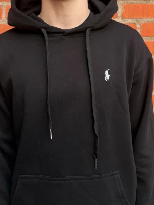 Svart hoodie från Polo Ralph Lauren - Snygg svart hoodie från Polo Ralph Lauren med klassisk huva och dragsnören. Liten vit broderad logga på bröstet och stor magficka framtill. Tillverkad i mjukt material som känns skönt mot huden. Perfekt för en avslappnad och stilren look.