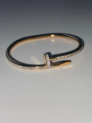 Guldfärgat armband med stenar - Snyggt armband i guldfärg format som en böjd spik, dekorerat med gnistrande små stenar längs hela ovansidan. Stilren och modern design som sticker ut och passar perfekt till dig som gillar unika accessoarer.