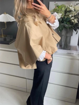 Kort beige trenchcoat med knappar - Säljer en trendig kort trenchcoat i beige med oversized passform och stora knappar framtill. Jackan har klassisk krage och är tillverkad i ett slätt bomullsliknande material. Perfekt att slänga över en stickad tröja för en cool lager-på-lager-look.