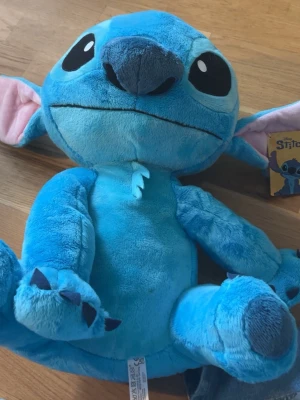 Stitch mjukisdjur Disney blå - Stor mjukisdjur av Stitch från Disney, i en klarblå färg med stora svarta ögon och rosa insida på öronen. Tillverkad i mjukt plyschmaterial och har detaljer som klor på tassarna och en liten tagg på bröstet. Perfekt för alla som älskar Lilo & Stitch!